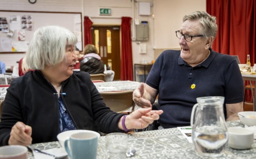 An older woman and an older man having a conversation while sat a table in a village hall